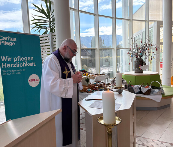 Bewohner*innen sitzen im Gemeinschaftsraum und nehmen an einer österlichen Messe mit Palmzweigen teil.