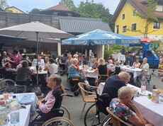 im Innenhof des Pflegewohnhauses sitzen zahlreiche Bewohnerinnen und Bewohner an festlich gedeckten Tischen, während im Hintergrund für Musik und Unterhaltung gesorgt wird.