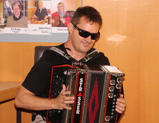 Ein blinder Mann sitzt auf einem Stuhl und spielt auf einer Ziehharmonika in den Farben Schwarz, Rot und Gold. Er trägt eine schwarze Sonnenbrille und wirkt konzentriert und zugleich entspannt beim Musizieren.