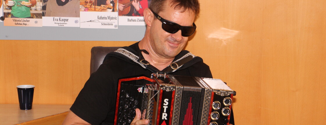 Ein blinder Mann sitzt auf einem Stuhl und spielt auf einer Ziehharmonika in den Farben Schwarz, Rot und Gold. Er trägt eine schwarze Sonnenbrille und wirkt konzentriert und zugleich entspannt beim Musizieren.