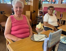 Zwei Bewohnerinnen sitzen gemeinsam im Café vor ihren Eisbechern und freuen sich darauf, das leckere Eis zu genießen.