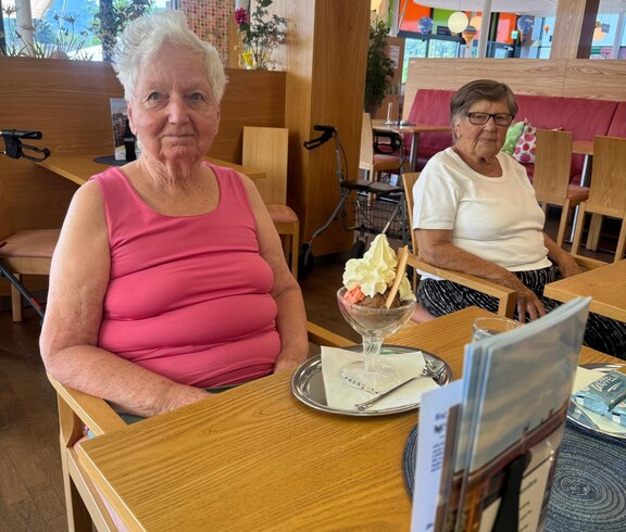 Zwei Bewohnerinnen sitzen gemeinsam im Café vor ihren Eisbechern und freuen sich darauf, das leckere Eis zu genießen.