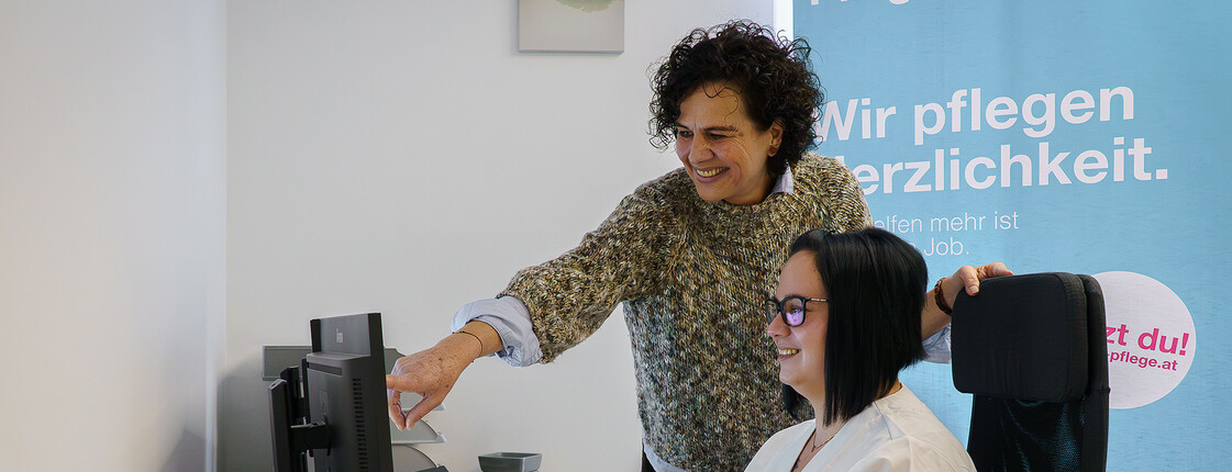 Zwei Caritas Mitarbeiterinnen arbeiten gemeinsam am Computer unter Banner „Caritas Pflege – Wir pflegen Herzlichkeit“