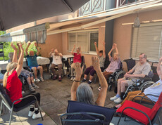 Bewohner*innen sitzen im Kreis auf der Terrasse und bewegen Arme zur Musik bei einer Bewegungsrunde im Freien.