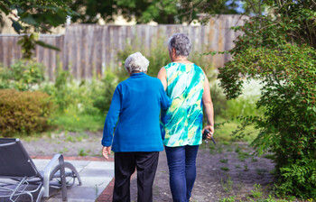 Zwei Damen gehen miteinander spazieren, wobei eine sich bei der anderen einhackt. Beide sieht man nur von hinten.