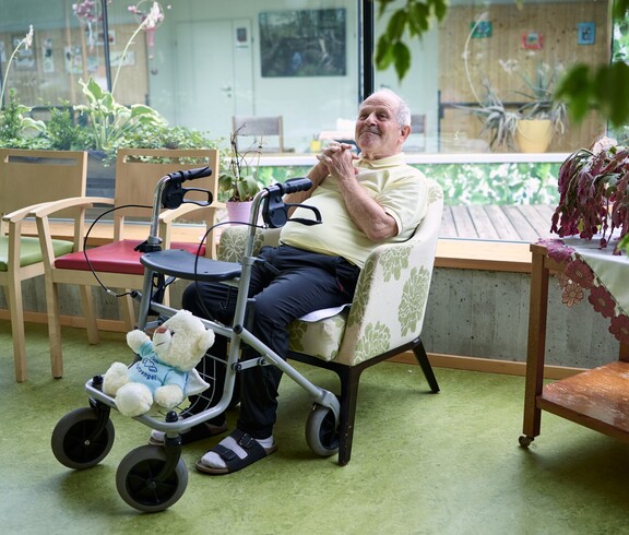 Ein älterer Herr sitzt entspannt in einem Sessel in einem hellen Aufenthaltsbereich eines Pflegewohnhauses. Vor ihm steht ein Rollator, in dessen Korb ein Stofftier sitzt. Neben ihm stehen bunte Stühle, und durch die große Fensterfront fällt Tageslicht auf zahlreiche Pflanzen, die den Raum freundlich und grün wirken lassen.