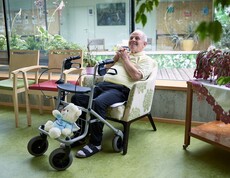 Ein älterer Herr sitzt entspannt in einem Sessel in einem hellen Aufenthaltsbereich eines Pflegewohnhauses. Vor ihm steht ein Rollator, in dessen Korb ein Stofftier sitzt. Neben ihm stehen bunte Stühle, und durch die große Fensterfront fällt Tageslicht auf zahlreiche Pflanzen, die den Raum freundlich und grün wirken lassen.