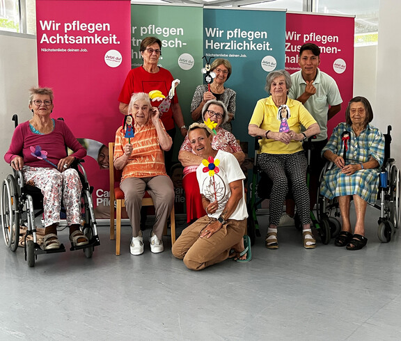 Senior*innen und Pfleger*innen präsentieren gebastelte Figuren vor bunten Caritas-Bannern.