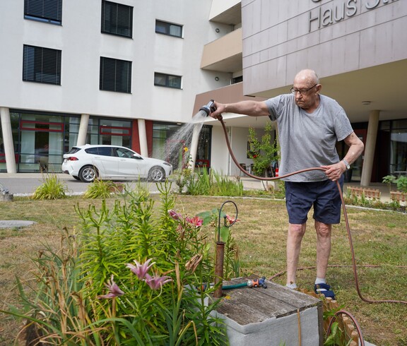 Ein Bewohner gießt mit dem Gartenschlauch die Pflanzen vor dem Eingang der Einrichtung.