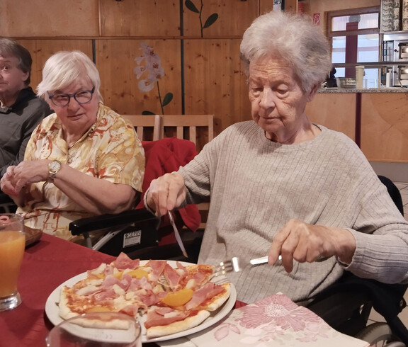Seniorin im Rollstuhl schneidet Pizza, andere Personen essen gemeinsam in einem Lokal