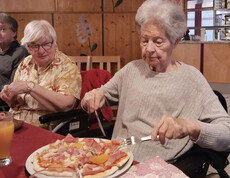 Seniorin im Rollstuhl schneidet Pizza, andere Personen essen gemeinsam in einem Lokal