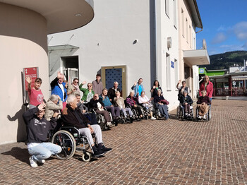 Große Gruppe von Senior*innen und Begleitpersonen winkt lachend vor dem Bahnhof Wolfsberg in die Kamera