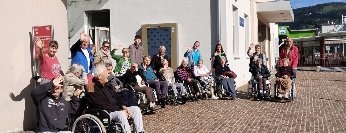 Große Gruppe von Senior*innen und Begleitpersonen winkt lachend vor dem Bahnhof Wolfsberg in die Kamera