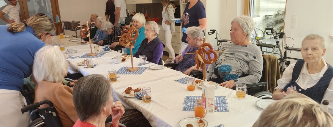 Oktoberfest-Tafel mit Bewohner*innen  Bewohner*innen sitzen gemeinsam an einer festlich gedeckten Tafel mit Brezen und Saft.