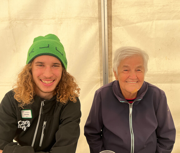 Zwei Personen sitzen nebeneinander und lächeln. Die Person links trägt eine grüne Mütze und eine schwarze Jacke. Die Person rechts hat kurze weiße Haare und trägt eine dunkle Jacke mit Reißverschluss. 