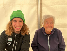 Zwei Personen sitzen nebeneinander und lächeln. Die Person links trägt eine grüne Mütze und eine schwarze Jacke. Die Person rechts hat kurze weiße Haare und trägt eine dunkle Jacke mit Reißverschluss. 