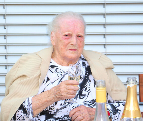 Frau Vacula sitzt an einem Tisch und hält ein Sektglas in der Hand, bereit zum Anstoßen mit ihrer Familie und dem Pflegepersonal.