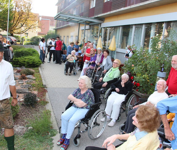 Bewohnerinnen und Bewohner des Caritas Pflegewohnhauses verfolgen vom Garten und von der Terrasse aus das Herbstfest, während ein Mitglied der Pischelsdorfer Schuhplattler in Tracht das Programm im Freien begleitet.