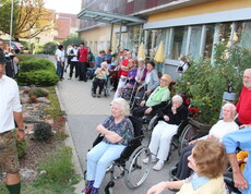 Bewohnerinnen und Bewohner des Caritas Pflegewohnhauses verfolgen vom Garten und von der Terrasse aus das Herbstfest, während ein Mitglied der Pischelsdorfer Schuhplattler in Tracht das Programm im Freien begleitet.