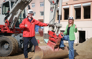 Zwei Personen mit Helmen stehen vor Bagger auf Baustelle und zeigen Daumen hoch im Kontext des Caritas Social Deals.