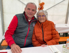 Zwei Personen sitzen an einem Tisch. Die Person links trägt eine graue und rote Jacke, die Person rechts eine orange Jacke.