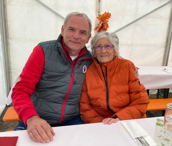 Zwei Personen sitzen an einem Tisch. Die Person links trägt eine graue und rote Jacke, die Person rechts eine orange Jacke.