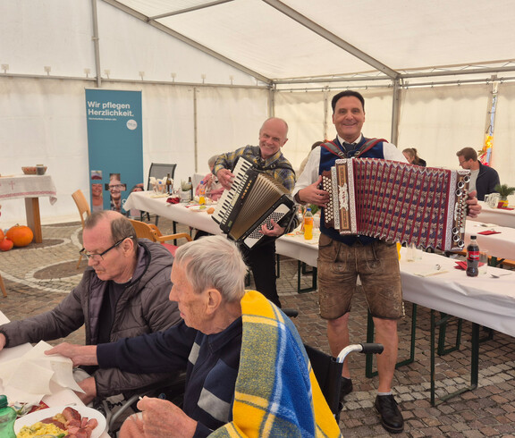 Zwei Musiker spielen Akkordeon im Zelt. Im Vordergrund sitzen zwei ältere Personen an einem Tisch mit Essen und Getränken. Ein Banner im Hintergrund trägt die Aufschrift "Wir pflegen Herzlichkeit".