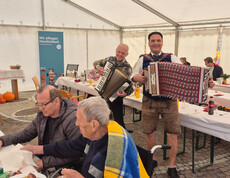 Zwei Musiker spielen Akkordeon im Zelt. Im Vordergrund sitzen zwei ältere Personen an einem Tisch mit Essen und Getränken. Ein Banner im Hintergrund trägt die Aufschrift "Wir pflegen Herzlichkeit".