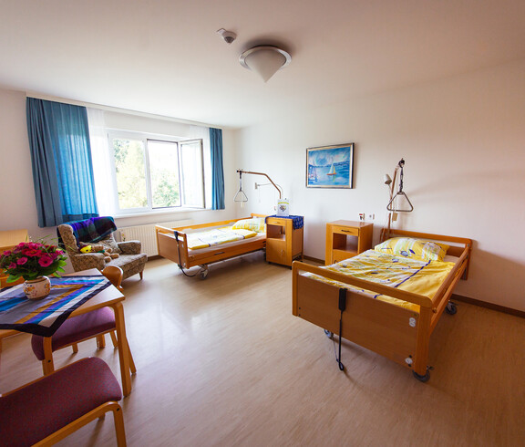 Ein Zimmer mit einem zwei Bett für pflegebedürftige Personen, 2 Nachtkästchen, 3 Sesseln, 1 Tisch mit Tischtuch und darauf stehender Blume sowie 1 Stuhl und 2 Bilder