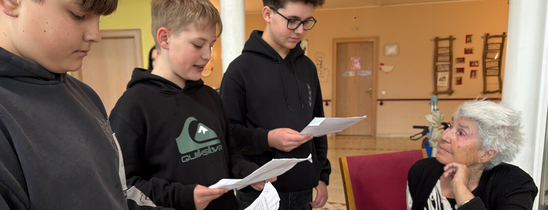 Schüler der Mittelschule Friesach lesen einer Bewohnerin im Gemeinschaftsraum von Haus Suavitas vor.