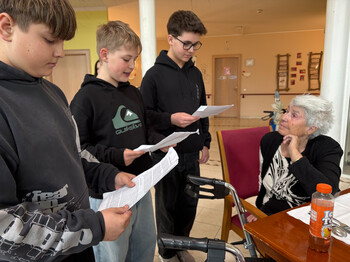 Schüler der Mittelschule Friesach lesen einer Bewohnerin im Gemeinschaftsraum von Haus Suavitas vor.