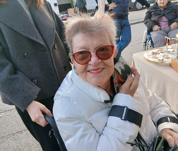 Person in weißer Jacke und roten Sonnenbrillen im Rollstuhl, lächelnd auf einem Markt.