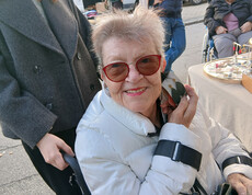 Person in weißer Jacke und roten Sonnenbrillen im Rollstuhl, lächelnd auf einem Markt.