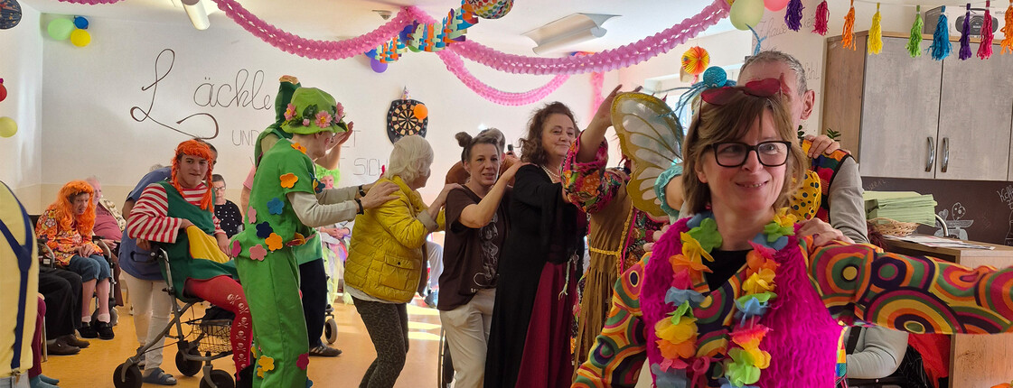 Bewohner*innen und Mitarbeitende in farbenfrohen Kostümen tanzen und feiern im geschmückten Gemeinschaftsraum.