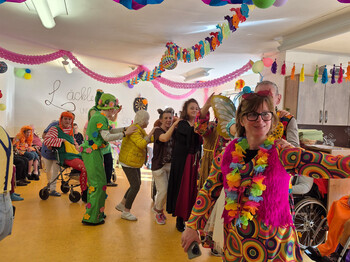 Bewohner*innen und Mitarbeitende in farbenfrohen Kostümen tanzen und feiern im geschmückten Gemeinschaftsraum.