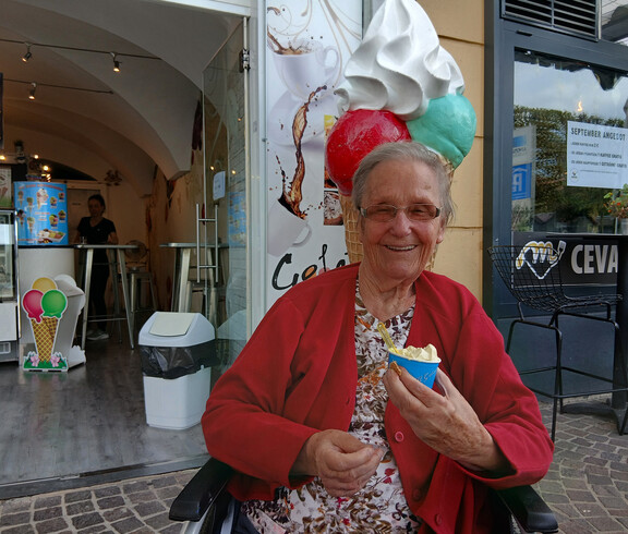 Seniorin im Rollstuhl sitzt vor einer Eisdiele und lacht mit einem Becher Eis in der Hand.