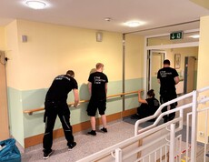 Vier Männer – mit dem Rücken zur Kamera – tragen Firmen-Shirts mit der Aufschrift „The Fortna Way“. Sie montieren einen langen Haltegriff entlang der frisch ausgemalten Wand.