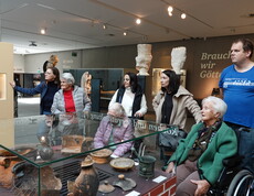 Mehrere Bewohner*innen in Rollstühlen stehen vor einer Vitrine, in der alte Tongefäße ausgestellt sind. Sie wirken aufmerksam und interessiert, da sie einer Führung lauschen. Neben ihnen befindet sich das Pflegepersonal.