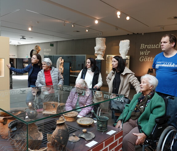 Mehrere Bewohner*innen in Rollstühlen stehen vor einer Vitrine, in der alte Tongefäße ausgestellt sind. Sie wirken aufmerksam und interessiert, da sie einer Führung lauschen. Neben ihnen befindet sich das Pflegepersonal.