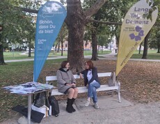 An einem Informationsstand zum Tag der Demenz sitzen zwei Personen auf einer Bank neben Broschüren und Informationsmaterialien, eingerahmt von zwei Fahnen in einer Parkanlage.