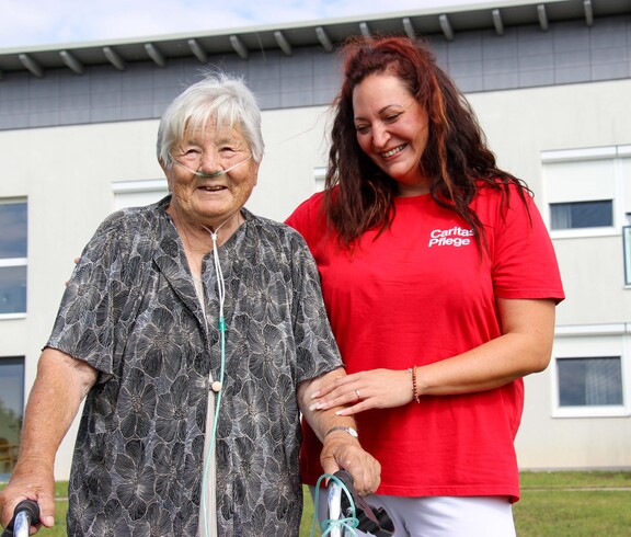 Eine Bewohnerin vom Haus Lisa, welche mit einem Gehwagen steht und Sauerstoffschläuche trägt. Neben ihr steht eine lächelnde Pflegekraft in einem roten T-Shirt mit der Aufschrift "Caritas Pflege", die sie unterstützend am Arm hält. Beide wirken zufrieden und fröhlich. 