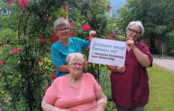 Drei Personen halten im Garten ein Schild mit der Einladung zum Aktionstag „Erinnern beugt Demenz vor“.