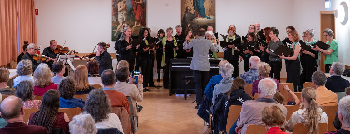 In einem Pfarrsaal sitzen viele Personen in Sitzreihen und hören einem Streichorchester am Ende des Raumes zu.