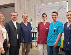 Hausleiterin Viktoria Trois (links) und Weihbischof Johannes Freitag (zweiter von links) stehen gemeinsam mit Mitarbeiterinnen des Caritas Pflegewohnhauses Eggersdorf vor dem Dienstplanbereich und posieren für ein Gruppenfoto.