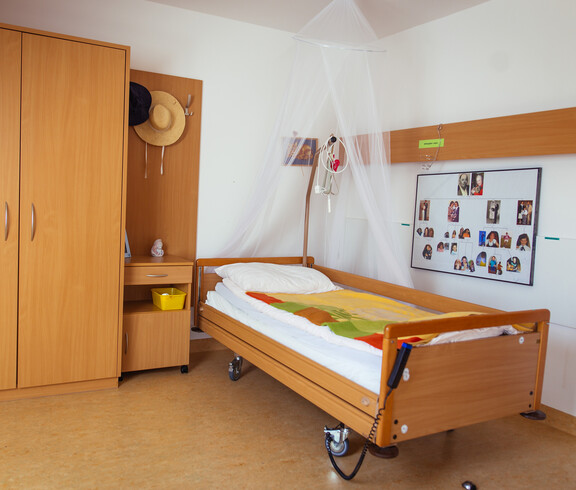 Ein Pflegebett steht an der Wand, auf der ein Bild mit kleinen Porträts hängt. daneben steht ein Nachschränkchen, dass mit dem Kleiderschrank verbaut ist.