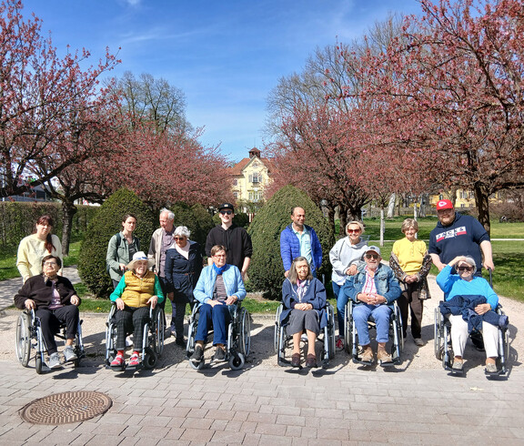 Gruppe von Menschen im Rollstuhl und Begleitpersonen pausiert unter blühenden Bäumen bei Frühlingsspaziergang in Klagenfurt.