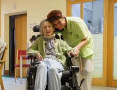 Pflegekraft beugt sich zu Rollstuhlnutzerin und hält ihre Hand