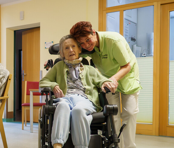 Pflegekraft beugt sich zu Rollstuhlnutzerin und hält ihre Hand