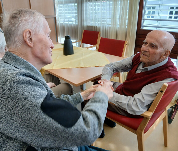 Zwei ältere Personen sitzen sich im Haus Magdalena gegenüber und reichen einander über einem Tisch die Hände.