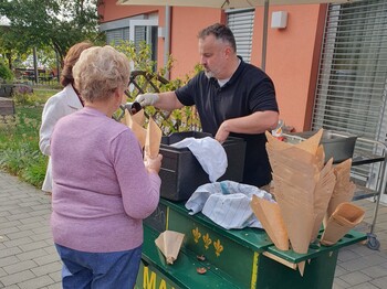 An einem Maronistand werden frisch gebratene Maroni in Papiertüten ausgegeben, während mehrere Personen im Freien auf der Terrasse zusammenkommen.